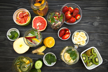Glasses of detox water with sliced fruit, vegetables, berries and herbs, set of ingredients: banana, strawberry, orange, grapefruit, kiwi, ginger, cucumber and herbs on black wood top view