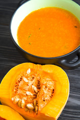 Pumpkin cream soup in a black ceramic bowl and pieces of fresh pumpkin on black wooden table