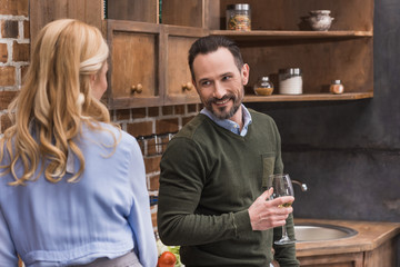 smiling husband with glass of wine talking to wife in kitchen