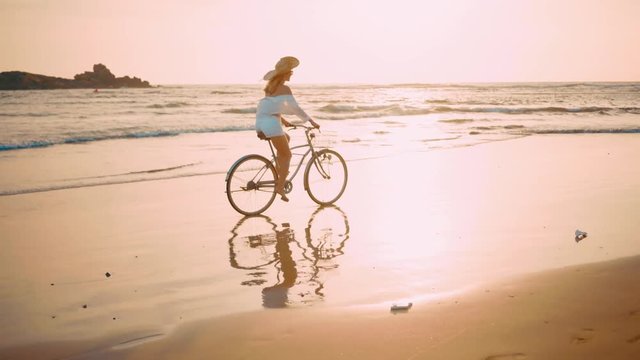 Woman in white and hat mooving on her bicycle along beach sand at summer time under burning sunset sky, lifestyle carefree.