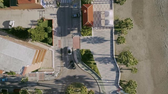 Bird's eye view aerial drone shot of simple and clean minimalistic suburban neighbourhood next to seaside and beach. Slow motion cars at roundabout, palm trees and calm.