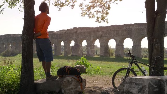 Cyclist with bike standing on a rock under a tree looking at ancient roman aqueduct. Young attractive athletic man with orange sportswear and backpack in parco degli acquedotti in Rome