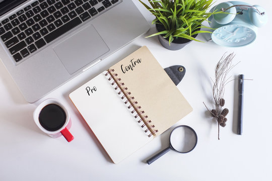 Laptop,spectacle,pen,dried flowers,coffee and a note book written with text PRO and CONTRA on white office table.Creative office table lay out.