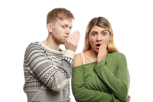 Attractive young unshaven male sharing secret or whispering gossips into his astonished girlfriend's ear, who is staring at camera with mouth wide opened, shocked with unexpected information