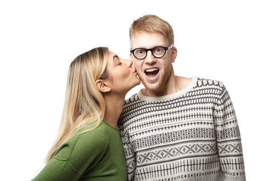 Studio shot of happy amazed young geeky guy with beard wearing eyeglasses and sweater opening mouth excitedly while being kissed by attractive blonde woman on cheek. Love and romance concept