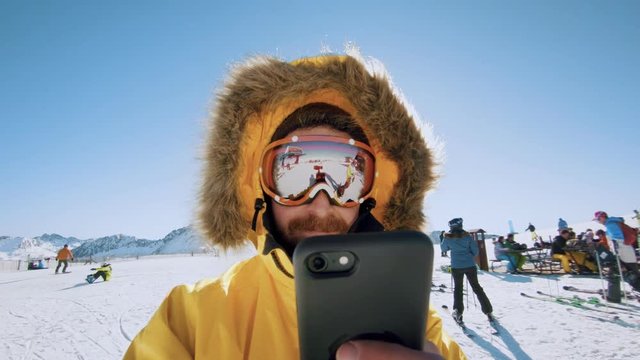 Funny and happy,optimistic man in active winter sports wear clothing, yellow jacket and hood,stands on top of mountain ski resort slope, makes video on action camera uses smartphone for selfie or text