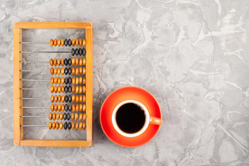 Old wooden scratched abacus near full red ceramic cup of black tasty coffee on round saucer on gray broken cement with crack. Top view