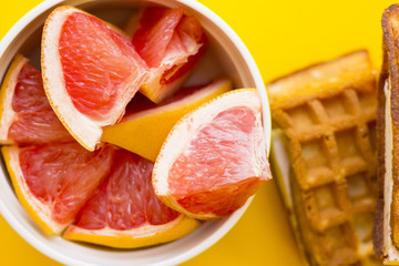 Naklejka na ściany i meble Delicious breakfast: fresh waffles and a bowl of sliced grapefruit on a yellow background with copy space, top view