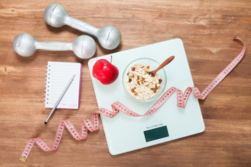Healthy diet, fitness and weight loss concept. Scales, measuring tape, oatmeal, apple, open notebook and pencil on the table. View from above.