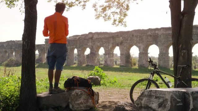 Cyclist with bike standing on a rock under a tree reading a map in front of ancient roman aqueduct. Young attractive athletic man with orange sportswear and backpack in parco degli acquedotti in Rome