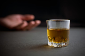 glass of whiskey or cognac or alcohol drink, hand of a drunk man in the background, alcoholism and alcohol abuse concept, defocused, selective focus, close up, gray table, dark background