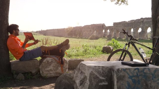 Cyclist with bike sitting on a rock under a tree on a videocall with his laptop in front of ancient roman aqueduct. Young attractive athletic man with orange sportswear and backpack in parco degli