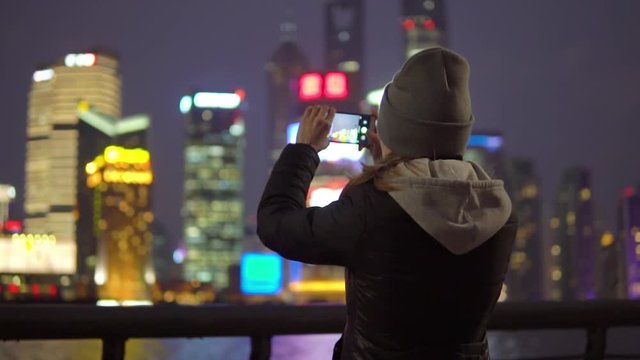 a young girl in a black jacket and hat takes pictures of Shanghai's attractions on mobile phone