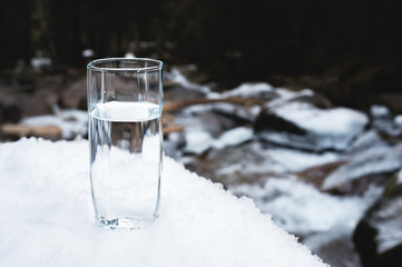 A transparent glass glass with drinking mountain water stands in the snow against a background of a clean mountain river and a forest in winter.