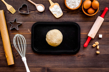 Ball of fresh raw dough near ingedients and cookware on dark wooden background top view
