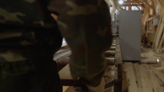 Caucasian carpenters puts wood board on refiner machine at lumber-mill, floor covered in sawdust