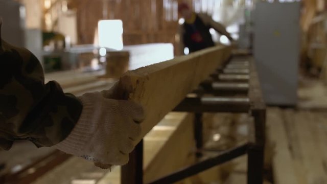Caucasian carpenters puts lumber plank on refiner machine at lumber-mill, floor covered in sawdust