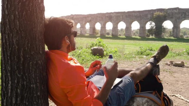 Cyclist with bike sitting on a rock under a tree listening to music with his laptop and drinking water in front of ancient roman aqueduct.