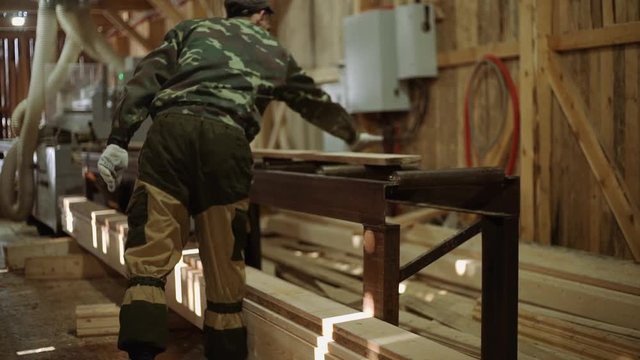 Caucasian carpenters puts wood plank on refiner machine at sawmill, floor covered in sawdust