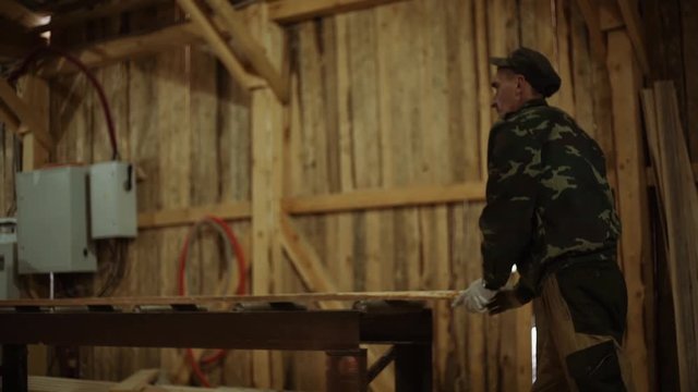 Caucasian carpenters puts lumber plank on refiner machine at sawmill, floor covered in sawdust