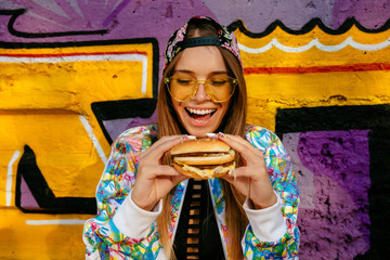Happy beautiful young woman, smiling widely, holds tasty burger in two hands. Dressed in bright...