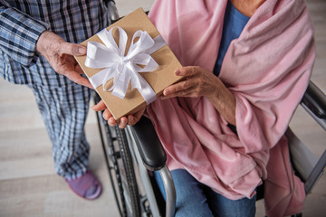 Top view close up of male and female hands holding the box with bow. Woman is sitting in invalid...