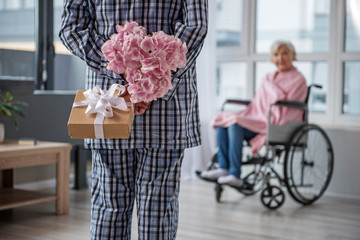 Husband standing in room with flowers and gift in his hands. He is hiding them behind his back....