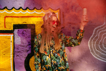 Attractive glad girl in sunglasses holds red smoke bomb, standing near the wall with graffiti. Dressed in colorful jacket and cap, with long hair.