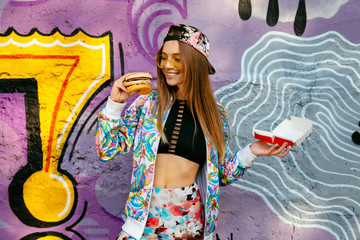 Funny beautiful girl in sunglasses holds a cheeseburger and open box of it. Dressed in colorful clothes, stylish cap, standing on the wall background with a graffiti in bright tones.