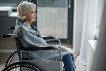 Moody senior woman looking outside with yearning look. She is sitting in wheelchair in room wrapped...