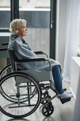 Cheerless senior female sitting in invalid chair with sad look. She is wrapped in a blanket and looking outside