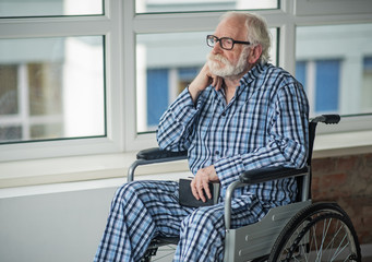Moody old man holding the book in his hands and sitting in invalid chair near the window. He is...