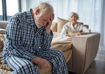 Feeling bad. Moody old husband sitting on bed with sad look and propping head with his hand. His...