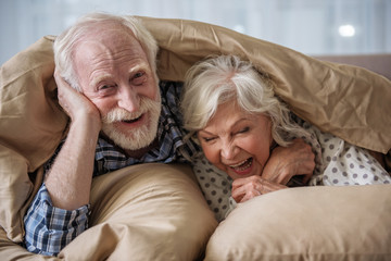 Cheerful old married couple lying in bed under blanket. Woman is laughing and man is looking at...