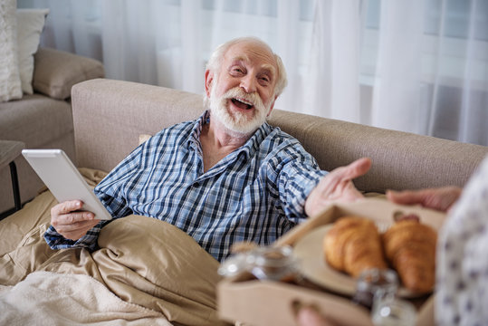 Focus on the face of cheerful senior man holding the device and sitting in bed while smiling. Old woman holding salver with breakfast