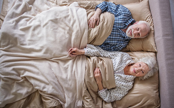 Top view of happy elderly husband and wife lying in bed. They are looking at camera with smile. Concept of peace