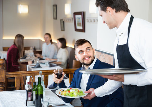 Waiter serving delicious salads to handsome young man at restaurant