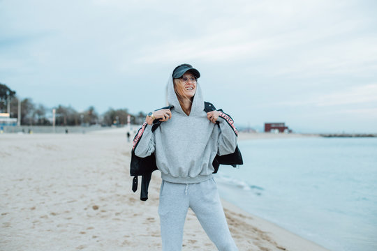 Happy and natural beauty young pretty and attractive woman or teenager wears grey hoodie and sweatpants during workout at beach seaside laughs naturally to camera, during fashion photoshoot influencer