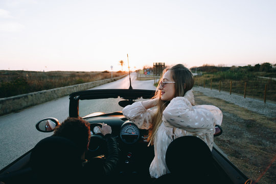 Excited and happy young female traveller or tourist enjoys warmth of summer inside rental convertible or cabriolet car on warm sunny day, palm trees aside road of holiday destination