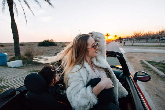 Happy and free, empowered young female woman or teenager stands up in seat of open roof cabriolet or convertible car. Raises her hands in warm air of summer vacation destination for bloggers