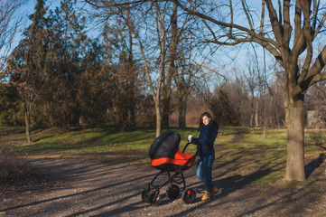 Young woman in blue coat walking in autumn park with little child baby in orange baby carriage, tea or coffe on fall nature, green trees. Mother and kid. Parenthood, love, parents, children concept.