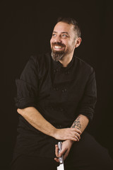 Portrait of a smiling chef looking to the left holding a knife on a black background.