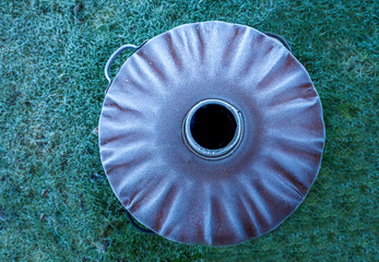 over head view point of the circular lid of a garden fire bin with a coating of white frost on it, a black circular whole in the middle, green grass background, the image has a lot of graphic texture