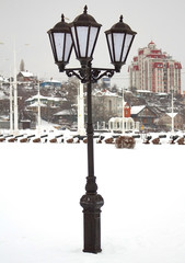 Iron, forged lantern, black color stands on the city street, on a winter day in the snow
