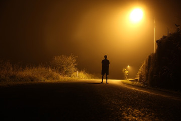 The silhouette of a man in shorts, standing in the middle of the road on a misty night. The glare of the street light against the fog sets a creepy mood. Concept of thieves, rapist and killers roaming - obrazy, fototapety, plakaty