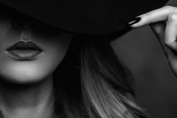 Beautiful young mysterious blond girl in black hat and black jacket on gray background. Eyes are covered with a hat. Manicure - long red nails, nail polish. Fashion, beauty.