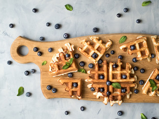 Traditional homemade belgian waffles with fresh ripe berries blueberry on wooden serving board over gray background. 
