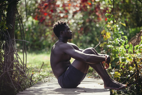 Side view of thoughtful shirtless man hugging knees while sitting on boardwalk