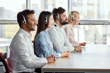 Side view happy multicultural call center operators sitting at desk. Call center workers with...