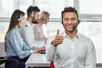 Cheerful american black operator with thumb up. Portrait of young male customer service representative, working co-workers background.